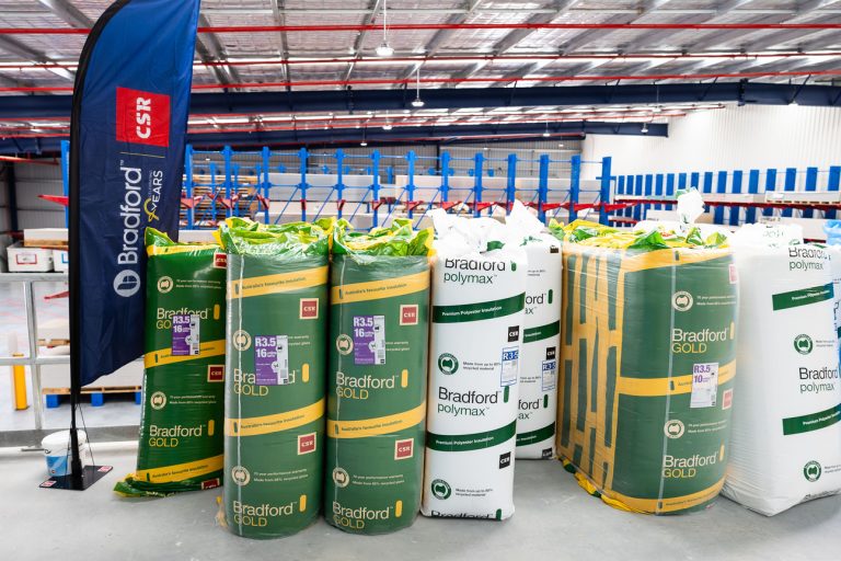 Stacks of Bradford Gold insulation rolls in green wraps and white Bradford polymer rolls in a warehouse, with a blue CSR-branded banner nearby.