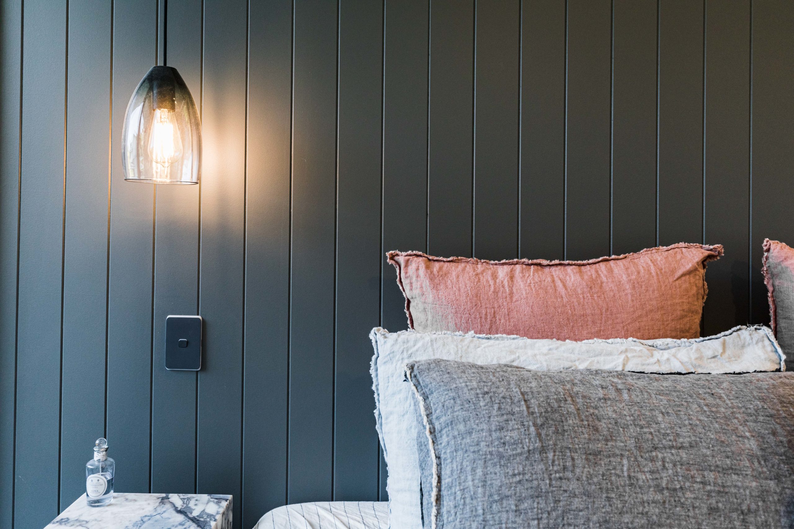 Warm glass pendant light casting a soft glow on a dark vertical-panel wall, with pink and gray cushions stacked on a sofa and a marble-topped table with a bottle nearby.
