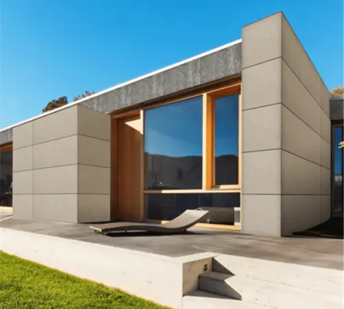 Modern single-story house with large glass doors and wooden-framed window, concrete block walls, a lounge chair on a concrete patio; green lawn nearby.