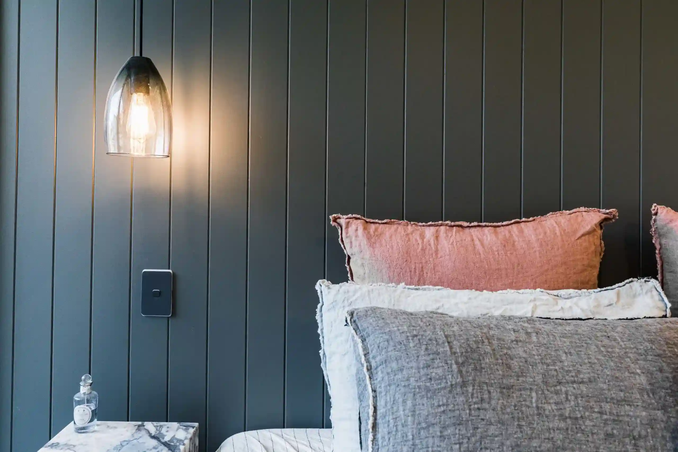 Dark vertical paneled wall with a hanging glass pendant light on the left, and pink and gray cushions along a sofa on the right.