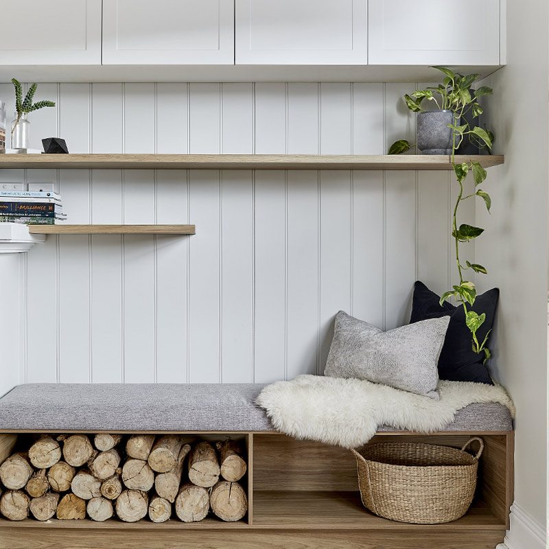Cozy bench with gray cushion, white fluffy throw, and pillows against a white paneled wall; under-seat storage filled with logs and a woven basket on the right.