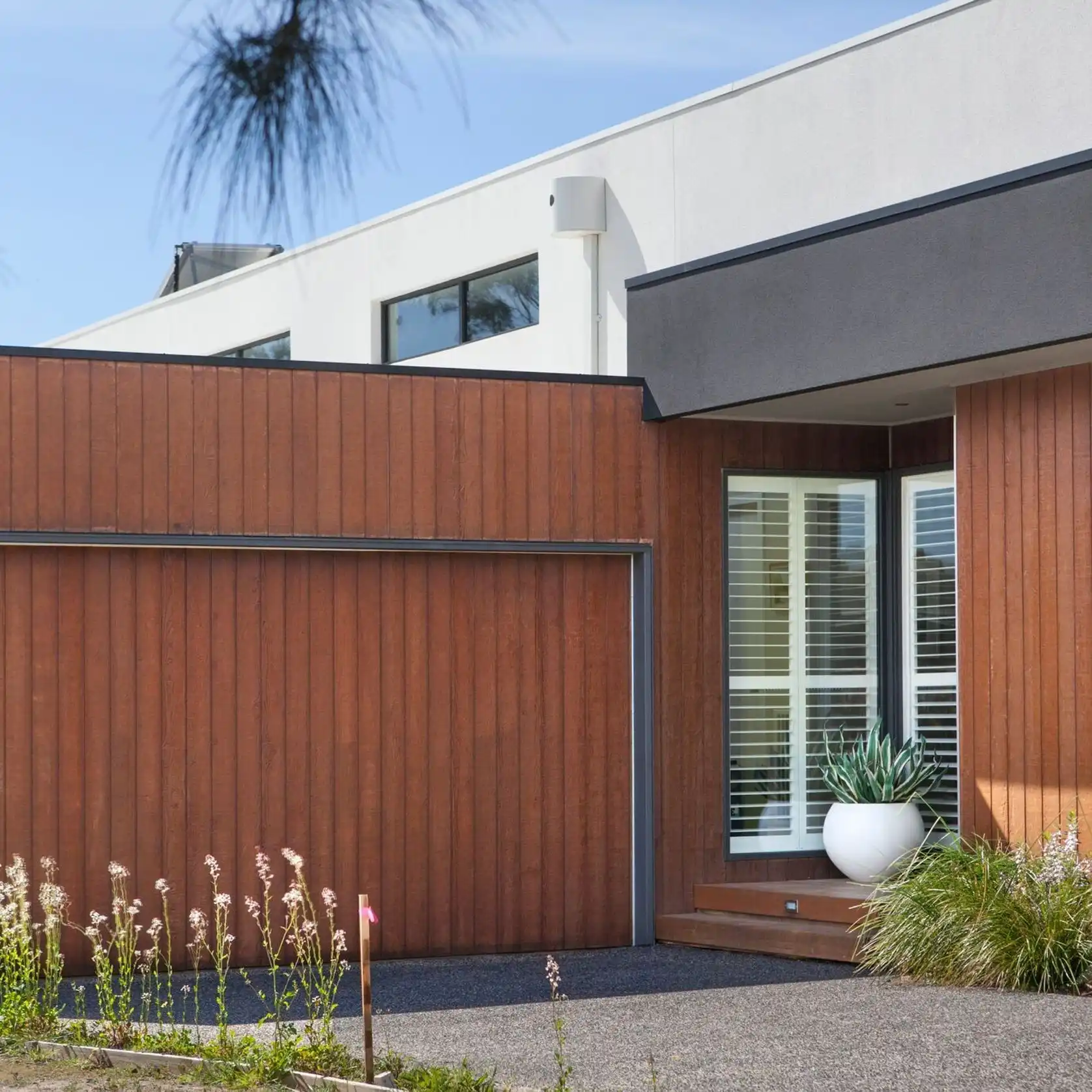 Contemporary house facade with vertical wooden panels, a white upper wall, and a large window with blinds near a wooden entry edge.