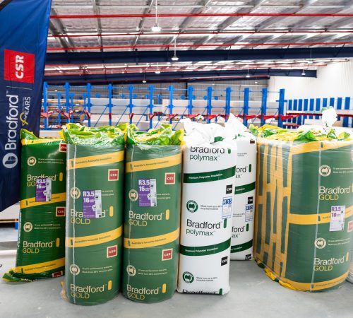 Stacks of Bradford Gold insulation rolls in green wraps and white Bradford polymer rolls in a warehouse, with a blue CSR-branded banner nearby.