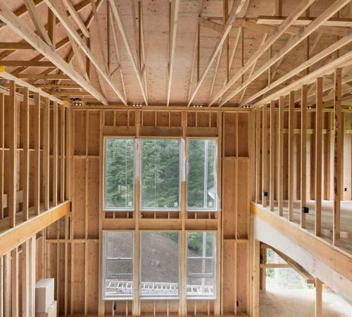Interior of a wooden-framed house under construction, with exposed studs and a large window revealing greenery outside